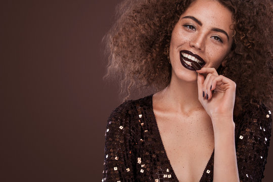 Beauty Portrait Of Young African American Girl With Afro Hairstyle And Chocolate . Girl Posing On Brown Background, Looking At Camera. Studio Shot.