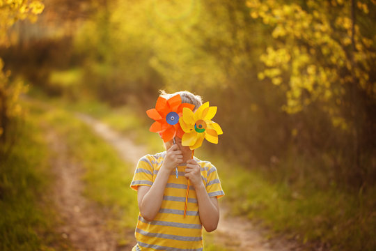Little boy hides behind yellow and orange pinwheels on green forest background in sunny day.