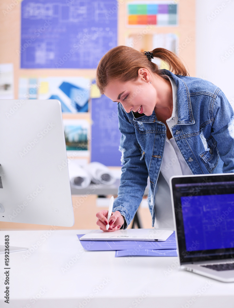 Wall mural young woman standing near desk with instruments, plan and laptop