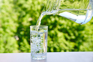  drinking water is poured from a jug into a glass on a green nature outdoors background . © evafesenuk