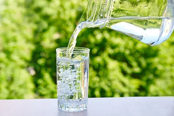 Fototapeten  drinking water is poured from a jug into a glass on a green nature outdoors background . © evafesenuk