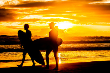 silhouettes of two surfboarders with boards walking by beach at beautiful golden sunset on sea beach background