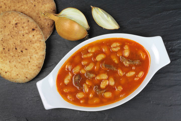 Traditional Turkish cuisine. Kuru Fasulye . Haricot Beans with onion and rustic bread on black stone background.