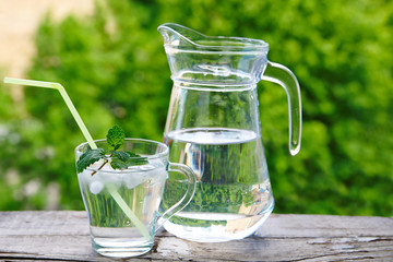 Glass with straws, ice water  and mint, a jug of water on a wooden table against a background of green foliage