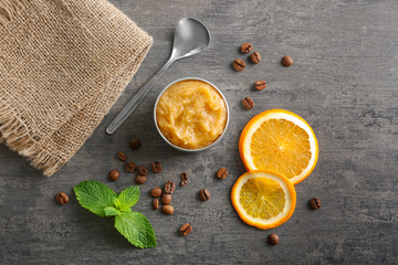 Jar with scrub, metal spoon, orange slices, coffee grains and mint on grey wooden background