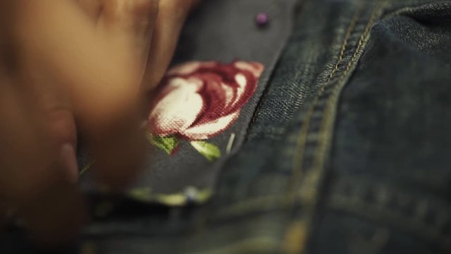 Unrecognizable female tailor hands stitch up cloth patch with rose image to denim jeans pocket in work shop close up slow motion
