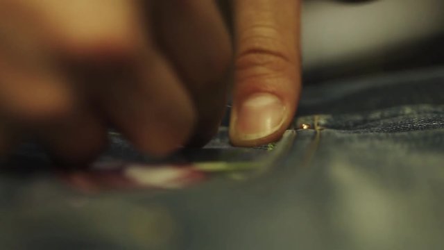 Unrecognizable woman tailor hands stitch up cloth patch with rose image to denim jeans pocket in work shop close up slow motion