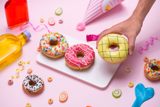 Party. Hand holding colourful sugary round glazed donuts and bottles of drinks on pink background.