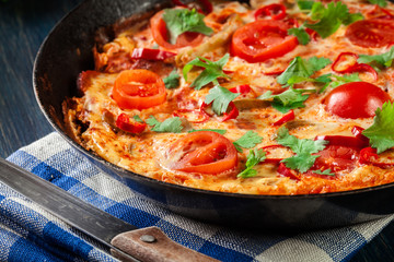 Frittata made of eggs, sausage chorizo, red pepper, green pepper, tomatoes, cheese and chili in a pan on wooden table