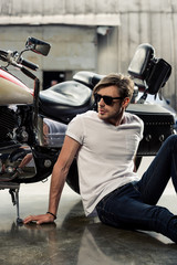 Fototapeta na wymiar Handsome stylish young man in sunglasses sitting near motorcycle and looking away