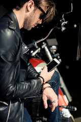 Side view of handsome stylish man in sunglasses and leather jacket using smartphone with blank screen