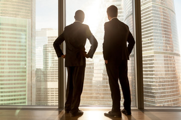 Rear view of african and caucasian businessmen wearing suits standing at full length large window...