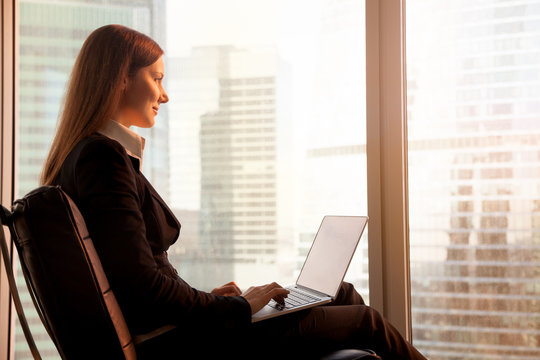 Side view of attractive businesswoman enjoying sunset, sitting comfortably in office chair with laptop on her knees, feeling inspired, calm and motivated for great achievements in career and life