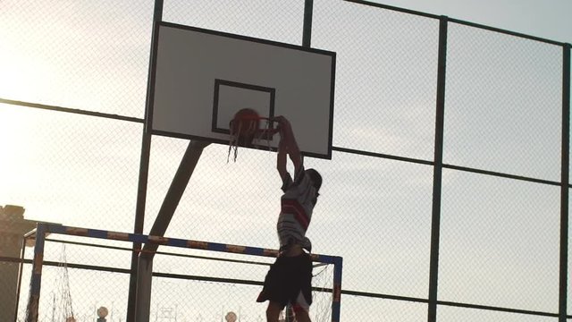 Caucasian male player in streetball doing two-handed slam dunk on outdoor sports ground in slowmotion