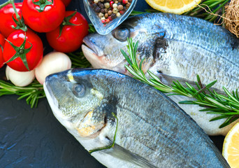 Fresh raw dorado fish with rosemary, garlic, tomatoes, pepper and lemon on a black slate background. Selective focus. Overhead view. Copy space.