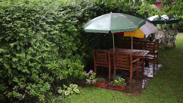 Summer rain sprinkles a garden sitting with parasols. Garden furniture in a green European style garden. 