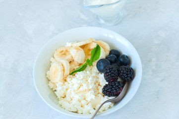 Homemade cottage cheese with bananas blueberries and blackberries, sour cream on a white stone background. Close up.