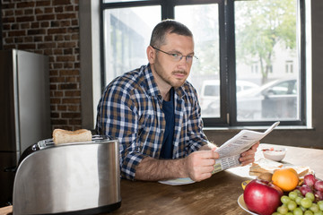 Serious bearded man in eyeglasses reading newspaper while sitting at table with fresh breakfast