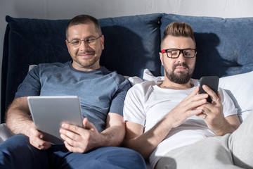Happy gay couple in eyeglasses lying together in bed and using digital devices