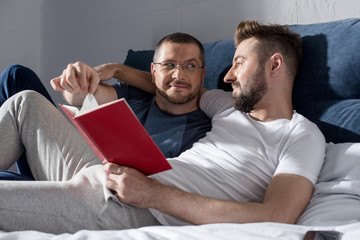 Happy homosexual couple lying together in bed and reading book