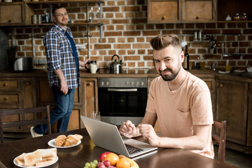 Smiling bearded man sitting at table and using laptop while partner pouring coffee behind
