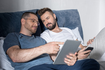 Happy homosexual couple using smartphone and digital tablet in bed