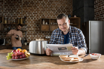 Smiling man in eyeglasses reading newspaper while bearded man pouring coffee behind