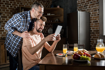 Smiling gay couple using digital tablet while having breakfast at home