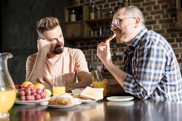 Bearded man looking at smiling partner in eyeglasses eating breakfast at home