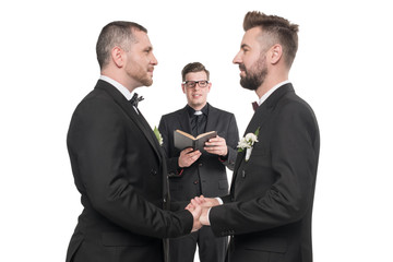 young homosexual couple in tuxedos holding hands at wedding ceremony