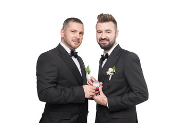smiling homosexual wedding couple in suits holding wedding rings and looking at camera