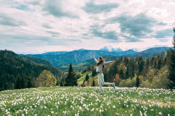 Fototapeta premium Young woman jumping in daffodil field, blooming narcissus with mountains on background
