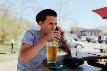 Handsome man in his 20s millennial sips on cold artisan craft beer ipa while talking on phone ordering a cab in summer outfit on open outside bar terrace