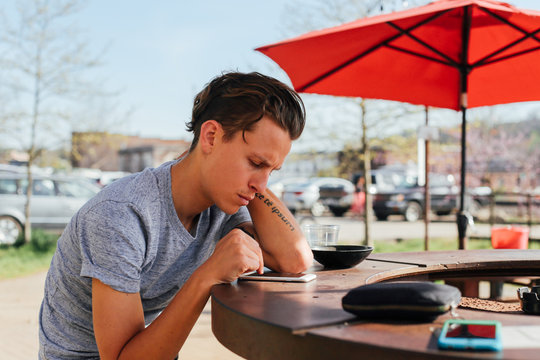 Handsome young man with stylish haircut and authentic tattoos waits for his car in outdoors cafe terrace, scrolls through his news feed on social media bored