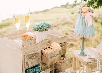 wedding decor outdoor. vintage beige dresser with small boxes decorated by sweets and fresh flowers. two glasses with champagne. nature recreation, date, engagement, romantic, event concept.