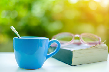 A blue cup of coffee with blurred eyeglasses, opened book and green natural background