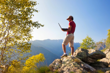 Young caucasian man sitting outdoor on a rock working on a laptop pc in mountain area.