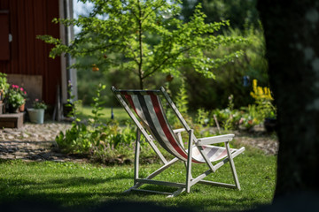 The sun shines from clear blue sky on the beautiful folding recliner made of wood and fabric, which is in the middle of the green and beautiful garden
