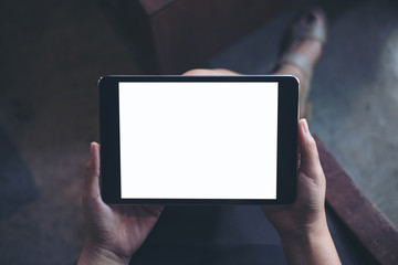 Mockup image of woman's hand holding black tablet pc with blank white screen on thigh with wooden floor background in modern cafe