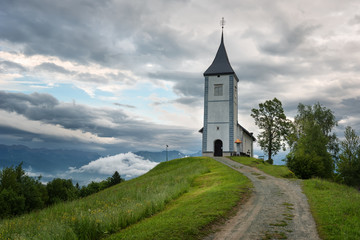 Fototapeta na wymiar Jamnik church on a hillside in the spring, foggy weather at sunset in Slovenia, Europe. Mountain landscape shortly after spring rain. Slovenian Alps. 