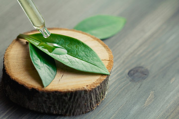 Close-up of cosmetic oil dripping from a pipette on a green leaf on a wooden cut.