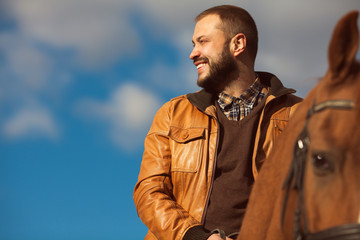 Country life concept. Young relaxed and smiling rich man in light brown leather jacket riding a horse. Sunny cloudy weather. Wide open spaces. Copy-space. Retro style. Close up. Outdoor shot