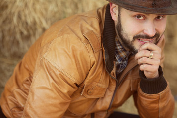Obraz premium Country life concept. Young relaxed and smiling rich man in light brown leather jacket sitting near straw bales. Close up. Outdoor shot