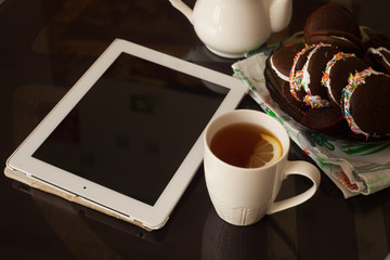 Cup of morning tea, teapot, chocolate cakes and tablet with blank screen on the table. front view. Morning blogger lifestyle background.