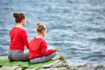 Rear view of young mother and daughter doing yoga exercises on the stone near the river.