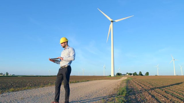 The engineer finishes work and leaves, looks at the windmills, holds a laptop in his hand.