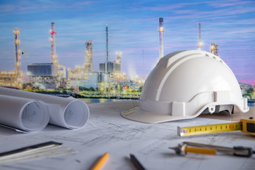 architectural drawing plan of refinery plant with blueprint rolls, protection safety helmet and project tools on work table, building construction industry concepts