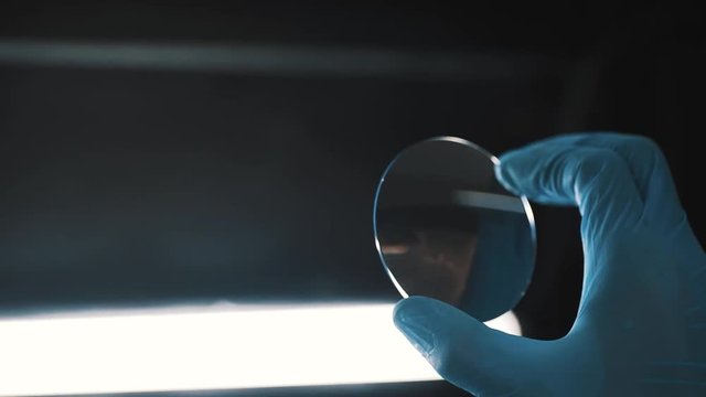 Scientist hand in blue rubber gloves checking round glass against white bright light in laboratory, close up
