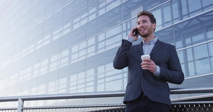 Businessman talking on mobile cell phone in city wearing suit in city business district. Confident young business man talking on smartphone. Urban male professional drinking coffee. SLOW MOTION.