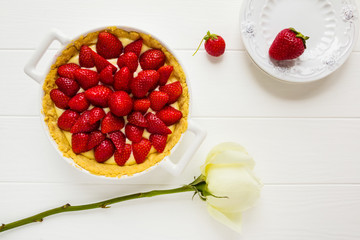 Strawberry custard tart in a white pan, vintage plates and a rose flower on the wooden table, top view.